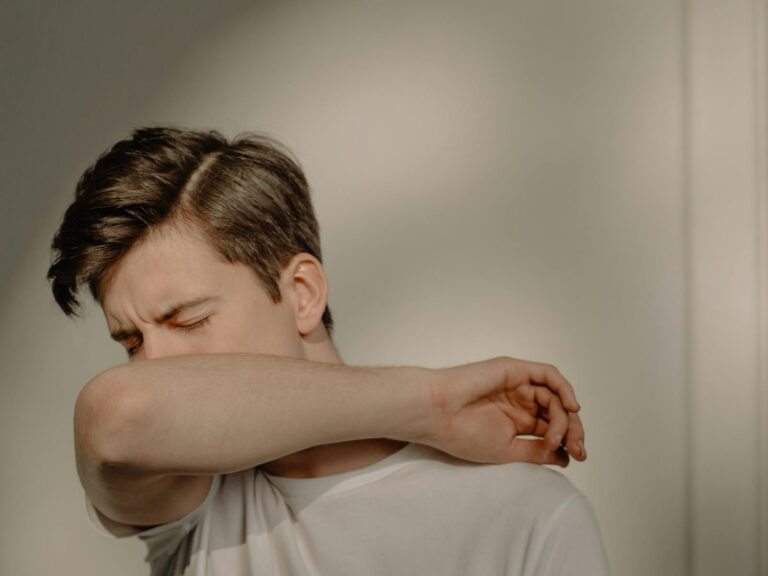 Pertussis (Keuchhusten) A young man sneezes into his elbow, showcasing a hygiene practice, in an indoor setting.