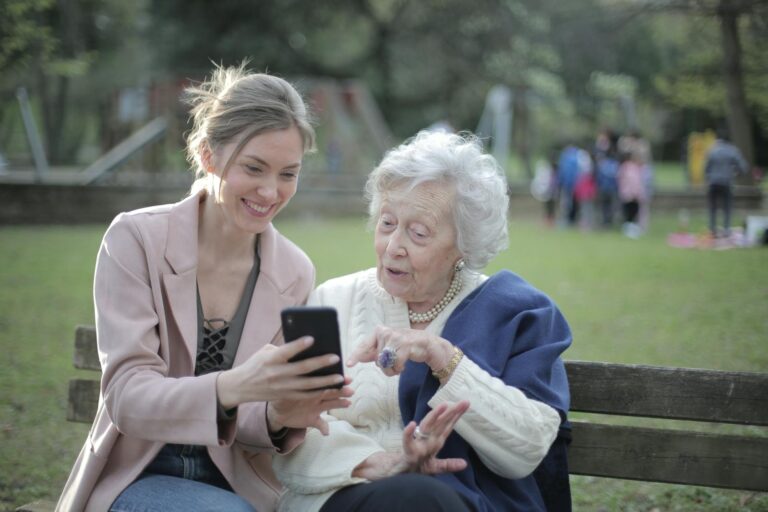 Alzheimer-Krankheit Delighted female relatives sitting together on wooden bench in park and browsing mobile phone while learning using