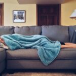 Person napping under a blue blanket on a comfortable couch in a modern living room setting.