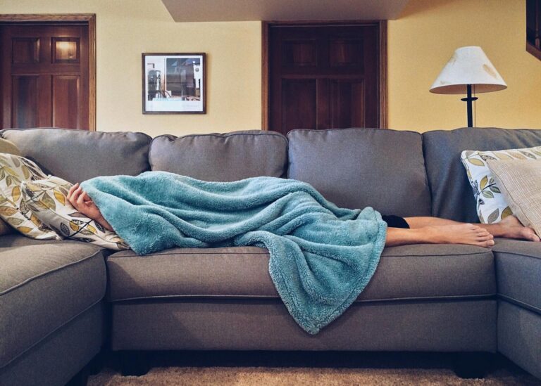 Katatonische Schizophrenie Person napping under a blue blanket on a comfortable couch in a modern living room setting.