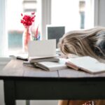 Blonde woman slumped over desk filled with books, showcasing study fatigue indoors.