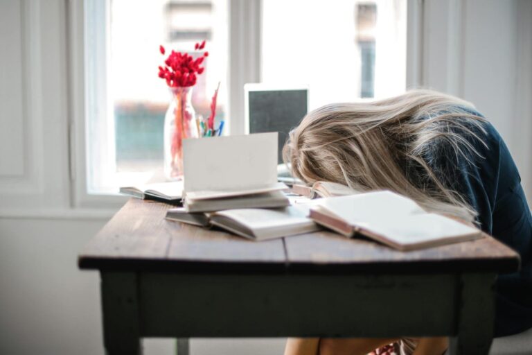 Nicht-organische Störung des Schlaf-Wach-Rhythmus Blonde woman slumped over desk filled with books, showcasing study fatigue indoors.