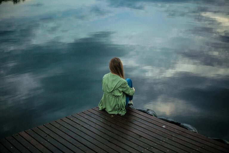 Abhängige (asthenische) Persönlichkeitsstörung A woman sits on a wooden dock, reflecting by a calm lake under a cloudy sky.
