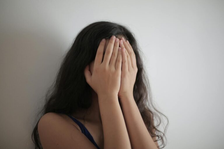 Vermeidende Persönlichkeitsstörung An adult woman with long brunette hair covers her face with her hands against a white wall, creating a minimalist portrait.