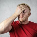 A frustrated man in a red shirt holds his head in stress against a neutral background.