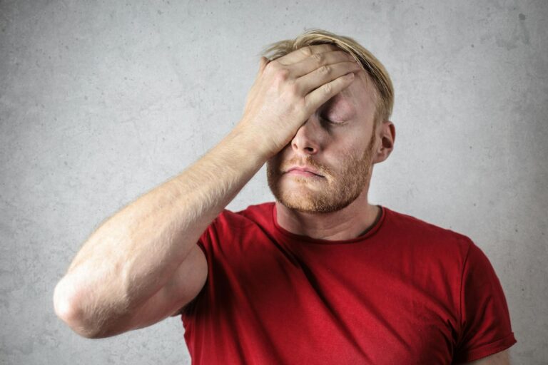 Atypische Gesichtsschmerzen A frustrated man in a red shirt holds his head in stress against a neutral background.