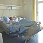 A woman lying in a hospital bed in a bright, softly lit room, appearing to rest or recover.