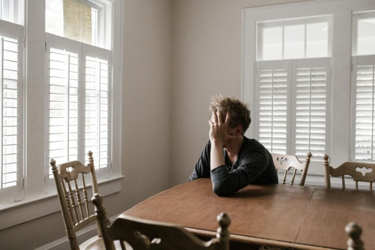 Somatoforme Störungen A man sits alone at a table in a bright room, displaying deep contemplation.